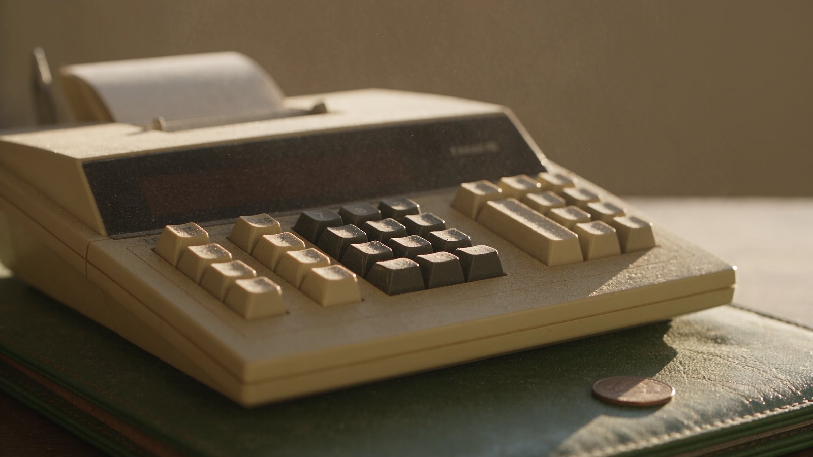 A dusty desk calculator on a leather blotter with a single coin beside it.