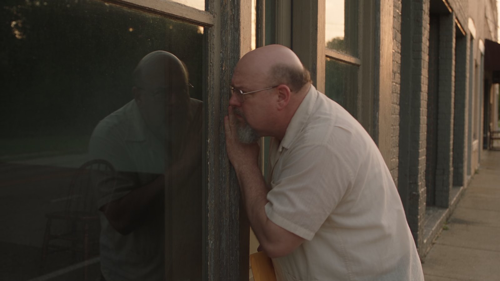 A man holding a yellow envelope knocks on a storefront door, then leans in to look through the glass at an empty room.