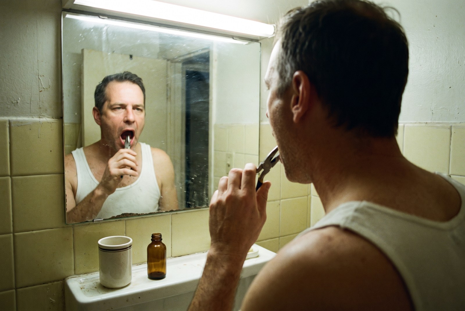 A man at a bathroom mirror reaching into his mouth with a pair of pliers.